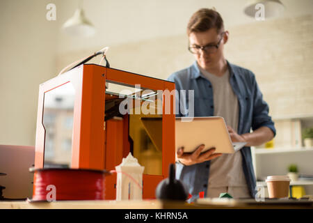 Concentrated young technician in eyeglasses using laptop while working on creation of 3D prototype, interior of modern laboratory on background Stock Photo