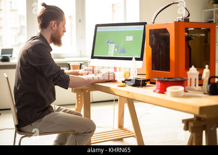 Profile view of confident bearded designer using computer program in order to create prototype with help of 3D printer, interior of modern studio on b Stock Photo