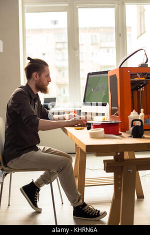 Full length portrait of concentrated young designer sitting in front of modern computer and prototyping 3D model, interior of spacious studio on backg Stock Photo
