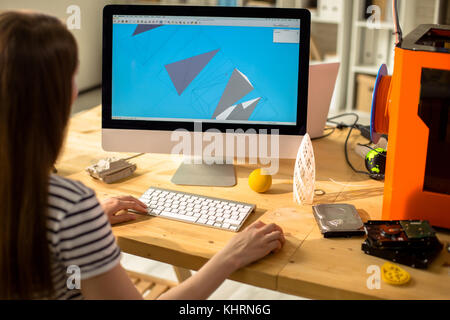 Over shoulder view of unrecognizable female designer using computer program in order to create detail drawing, 3D printer located on desk Stock Photo