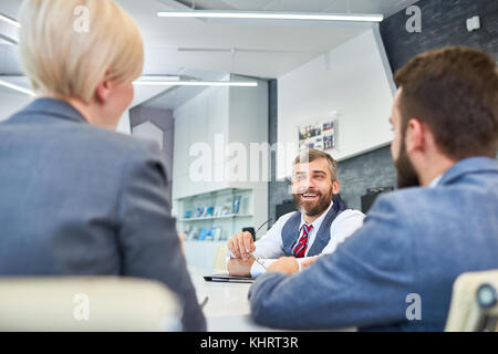 Portrait of barded mature businessman talking to partners and smiling while sitting at meeting table in board room while negotiating deal Stock Photo