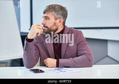 Profile view portrait of elegant mature man with greyish hair  sitting at meeting table in office listening to work discussion Stock Photo