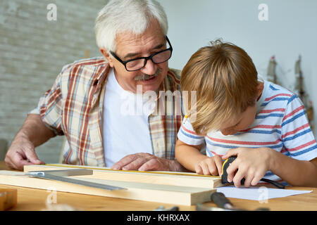 Portrait of old man teaching cute little boy woodwork, making wooden models together working at desk in small studio Stock Photo