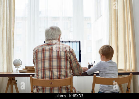 Rear view portrait of grandfather and grandson doing homework together sitting at desk with  blank screen computer Stock Photo