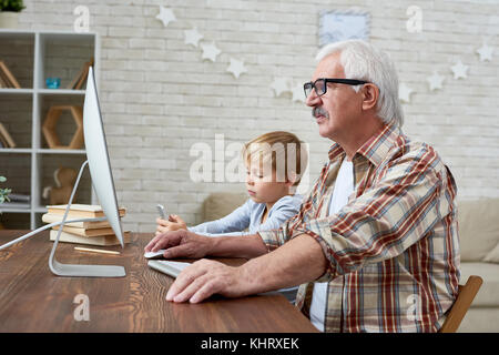 Portrait of senior man learning to use internet with tired little boy sitting next to him at desk at home Stock Photo