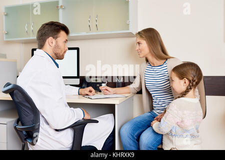 Portrait of little girl visiting doctor with mother, sitting at desk in office and consulting pediatrician for health check Stock Photo