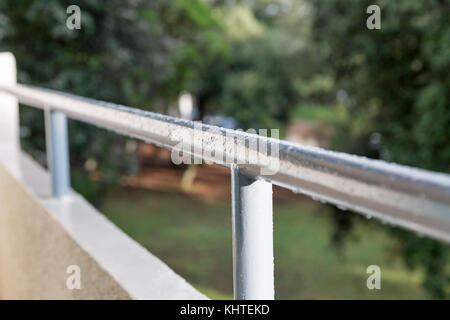 Raindrops on a metal balcony handrail closeup against a blurred background of green trees Stock Photo
