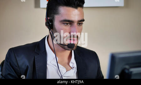 Good looking young man with headset working as help-desk assistant, customer service, help-line or telemarketer in office Stock Photo