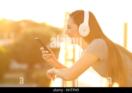 Side view portrait of a happy girl listening music with headphones and smart phone in a house balcony at sunset with an orange light Stock Photo