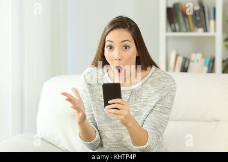 Front view portrait of a surprised woman holding a phone looking at you sitting on a sofa in the living room of a house interior Stock Photo