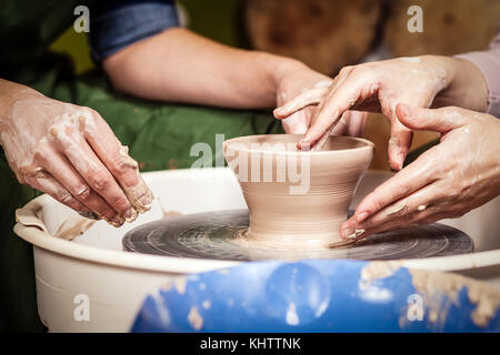 Close-up of a young woman potter is teaching a woman in a green apron to sculpt on a potter's wheel a vase of brown clay in a creative workshop, side  Stock Photo