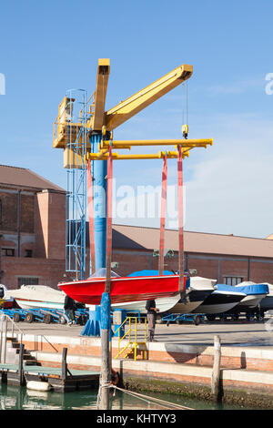Red boat with outboard motor suspended midair in slings on a boat lift or crane above the quay during launching at Giudecca, Venice, Italy in the boat Stock Photo