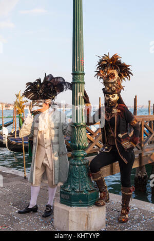 2017 Venice Carnival, Venice, Veneto, Italy, Two costumed people posing at the lagoon at sunset with gondolas Stock Photo