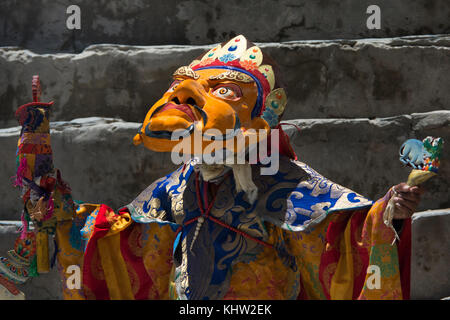 A Buddhist monk in an ancient masculine mask during a sacred ceremony in a monastery, in the hands of a wand and a drum. Stock Photo