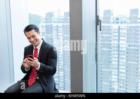 Businessman sitting on windowsill using smartphone smiling Stock Photo