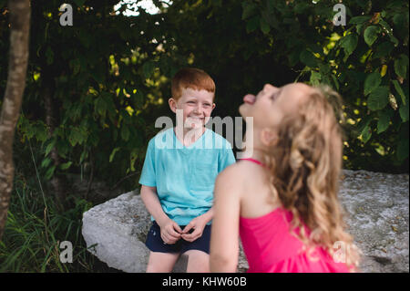 Girl sticking tongue out at brother sitting on rock Stock Photo