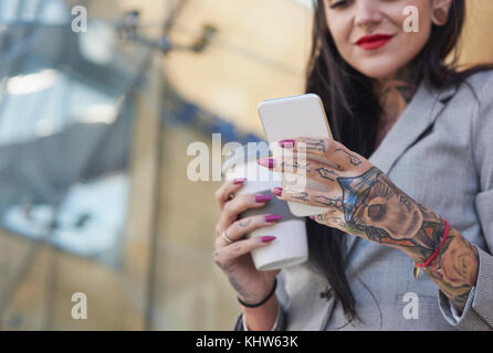 Businesswoman outdoors, holding coffee cup, using smartphone, tattoos on hands, mid section Stock Photo