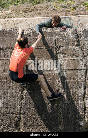 Young male free climber at top of sea wall helping friend climb up Stock Photo