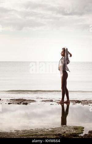 Young female runner at water's edge on beach stretching arms Stock Photo