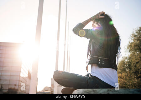 Young woman sitting cross-legged on wall, facing sunlight, tattoos on hand Stock Photo