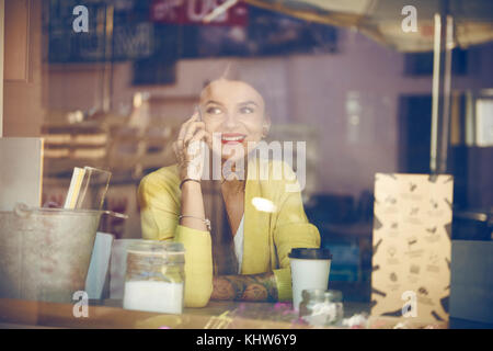 Young woman sitting in cafe, using smartphone, tattoos on hand, view through cafe window Stock Photo