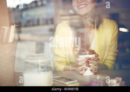 Young woman sitting in cafe, holding coffee cup, tattoos on hand, view through cafe window, mid section Stock Photo