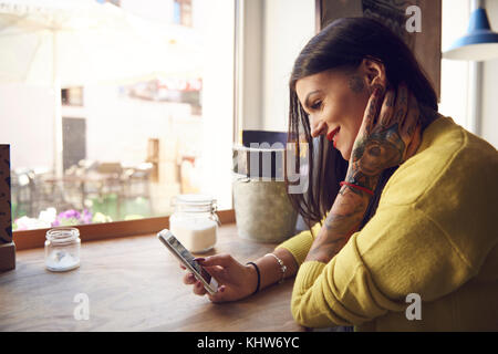 Young woman sitting in cafe, using smartphone, tattoos on arm and hand Stock Photo