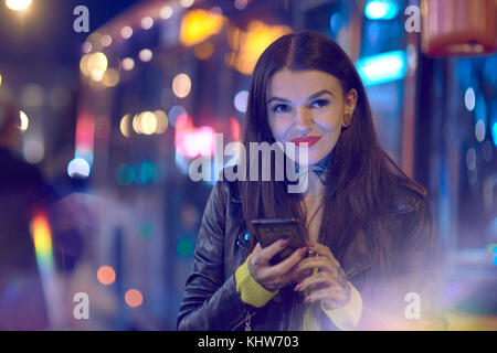 Young woman outdoors at night, holding smartphone, tattoos on hand and neck Stock Photo