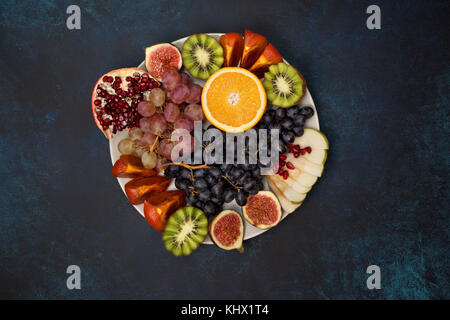 platter of fresh fruits: oranges, grapes, kiwi, persimmons, figs, pomegranate, apple on  blue background. view from above Stock Photo