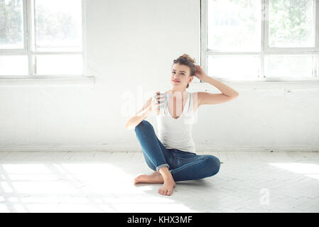 Attractive barefoot woman relaxing sitting on the floor holding a mug in her hand and smiling as she looks to the side in a high key room. Stock Photo