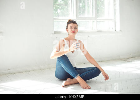 Attractive barefoot woman relaxing sitting on the floor holding a mug in her hand and smiling as she looks to the side in a high key room. Stock Photo