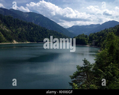 Peaceful freshwater lake at the Bavarian alps. Framed with the typical mountains and trees at a sunny day with blue sky and clouds. Stock Photo