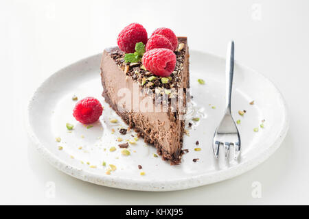 Slice of chocolate cake cheesecake decorated with crushed pistachio nuts, raspberries and mint leaf on white background. Closeup view Stock Photo