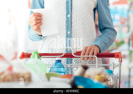 Woman doing grocery shopping at the supermarket, she is pushing a cart and checking a list Stock Photo