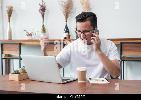 asia casual man arm up celebrate good new from mobile at laptop in coffee shop,success business concept,work outside office,work at home Stock Photo