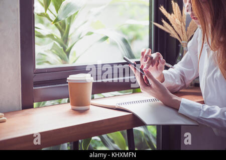 Businesswoman use mobile phone and writing a report on wood table at coffee shop near window,woking outside office lifestyle Stock Photo
