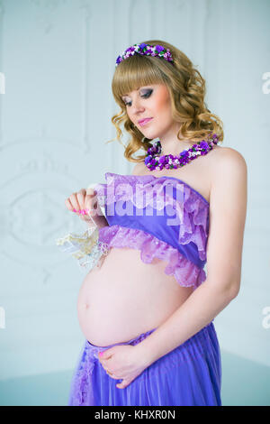 A happy pregnant woman in a purple long skirt poses in the studio against a background of flowers. Stock Photo