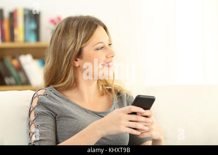 Girl imagining and holding a smart phone sitting on a sofa in the living room in a house interior Stock Photo