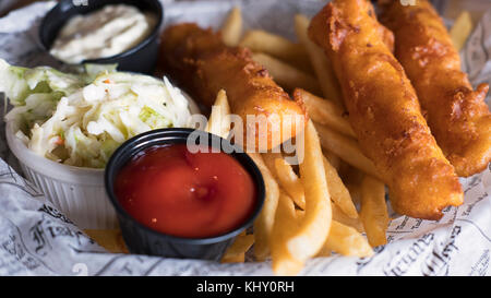 Tasty fish and chips, coleslaw and salsas served in English style on a plate covered with newspaper cuts Stock Photo