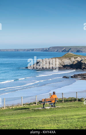 A man person wearing a bright red jacket sitting on a bench overlooking Trevelgue Head Porth Island in the background. Stock Photo