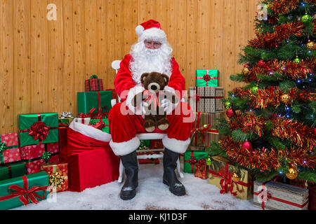 Santa Claus in his grotto surrounded by a Christmas tree with presents and gift wrapped boxes offering you a teddy bear. Stock Photo