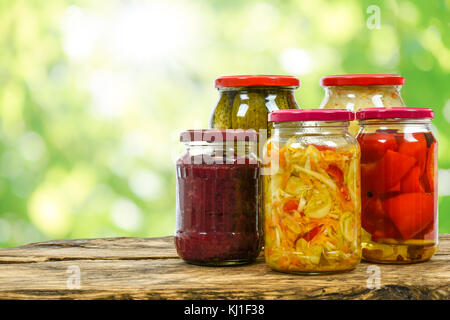 Composite image of jars with variety of homemade pickled vegetables on a wooden vintage table and on the blurred green garden background. Stock Photo