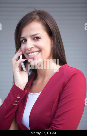 Portrait of young businesswoman in red blazer talking over the mobile phone in front of office building Stock Photo