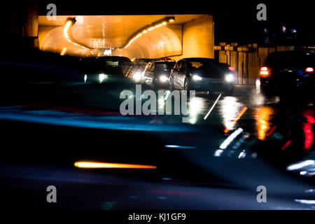 Night traffic on rainy city streets. Cars queued at tunnel exit waiting at intersection while driving vehicles moving past, front view with display da Stock Photo