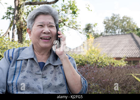 elder woman talk on mobile phone while sitting on bench in garden. elderly female speaking on smartphone in park. asian senior have phone conversation Stock Photo