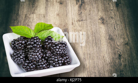 Sweet juicy blackberries with decorative green leaves in white square bowl. Vegetarian refreshment from luscious fresh fruit with a wooden background. Stock Photo
