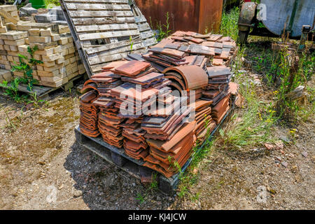 Old abandoned clay roof tiles lying on a pallet in an old yard.  Stack of bricks in the background. Stock Photo