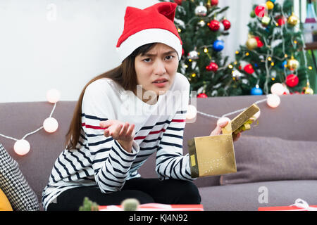 Asian woman upset when open gold xmas gift box at holiday party on sofa,boredom Christmas party present Stock Photo