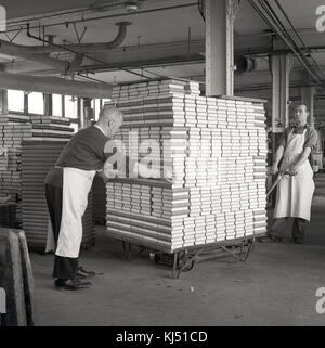 1950s, historical, two male workers wearing aprons inside an industrial unit or warehouse, moving a large and heavy load of hardback books on a platform trolley, England, UK. Stock Photo