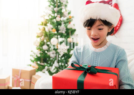little cute Asian girl opens her mouth feel shocked surprised to receive a big gift box for Christmas sitting on sofa of the living room at home. Stock Photo